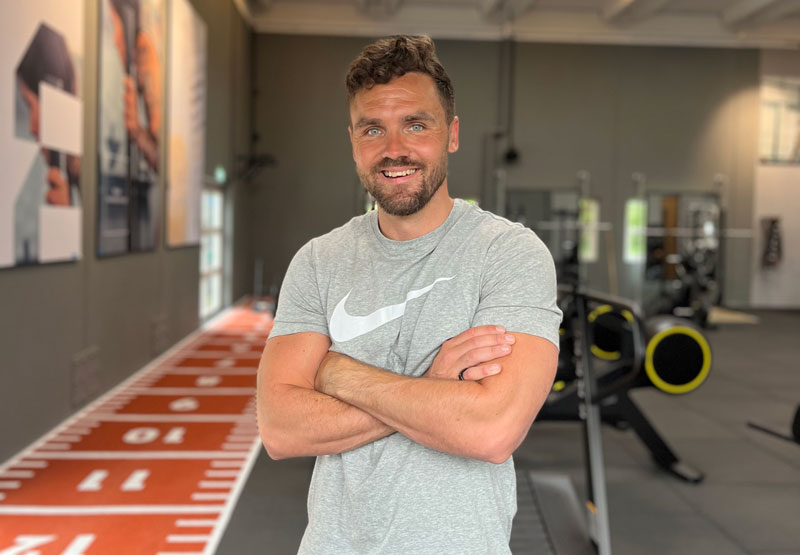 Et foto af træneren der står i et fitnesscenter og smiler.